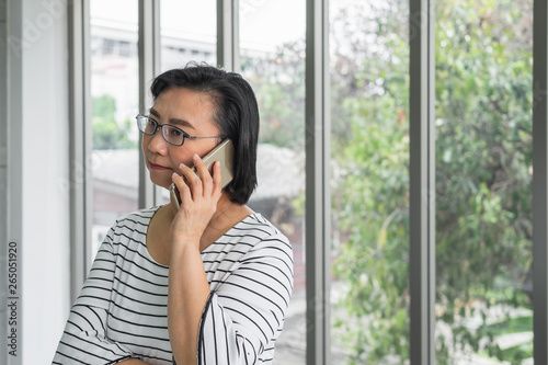 Billede på lærred An old Asian woman mother holding a phone in front of a window looking concern and serious