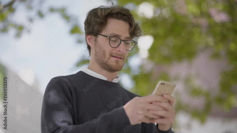 Smiling man in eyeglasses using smartphone. Low angle view of handsome happy man texting by cell phone outdoor, handheld shot. Technology concept Smiling man in eyeglasses using smartphone. Low angle view of handsome happy man texting by cell phone outdoor, handheld shot. Technology concept