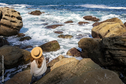 Girl in hat sittig on the rock and enjoying the view on the sea with white foam waves hittng rocks photo