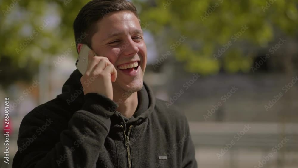Man talking by smartphone and laughing outdoor. Handheld shot of cheerful young man having pleasant phone conversation while standing on street at sunny day. Communication concept Man talking by smartphone and laughing outdoor. Handheld shot of cheerful young man having pleasant phone conversation while standing on street at sunny day. Communication concept