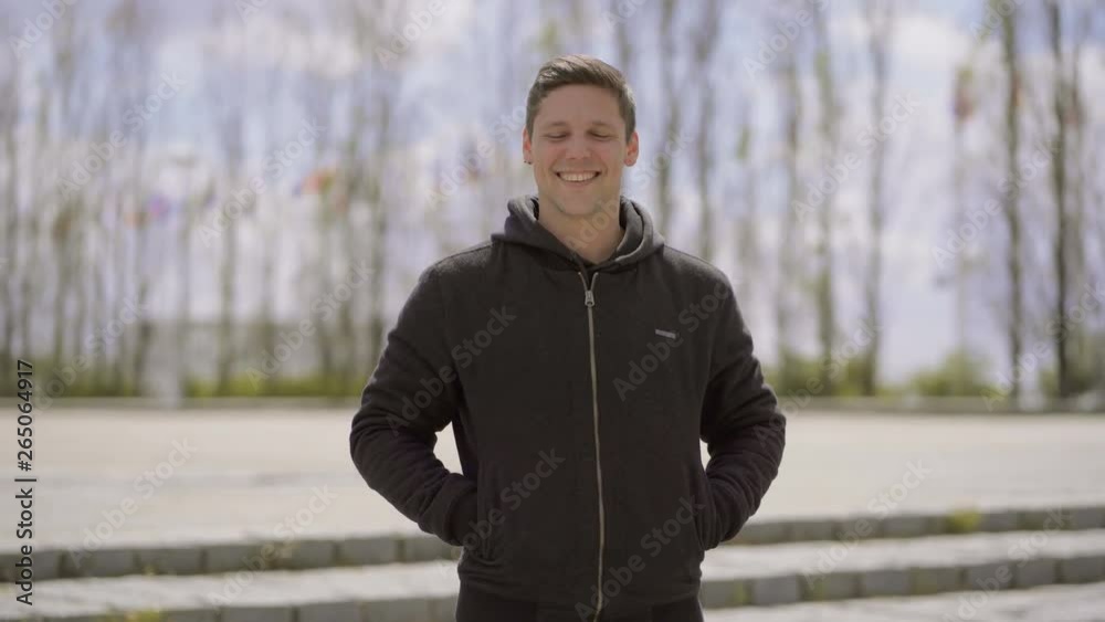 Handsome happy young man smiling at camera outdoor. Portrait of cheerful young man in hoodie standing with hands in pockets and crossed arms, laughing and looking at camera on street. Emotion concept Handsome happy young man smiling at camera outdoor. Portrait of cheerful young man in hoodie standing with hands in pockets and crossed arms, laughing and looking at camera on street. Emotion concept