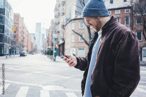 Young smiling man wearing hat and jacket walking on the city street typing on mobile phone in hand