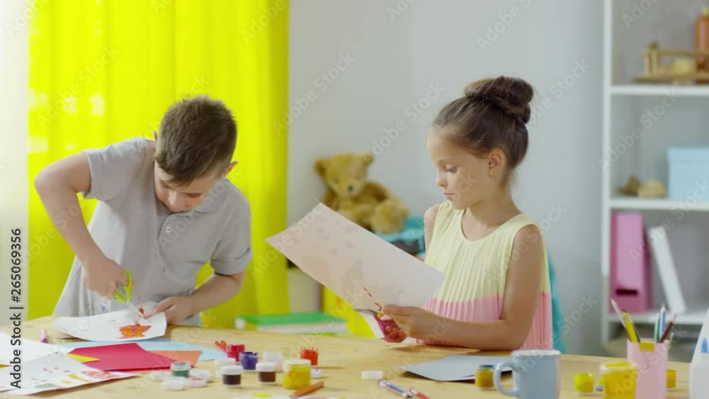 PAN of cute little girl and boy of preschool age cutting out paintings from paper and talking in art class in kindergarten PAN of cute little girl and boy of preschool age cutting out paintings from paper and talking in art class in kindergarten