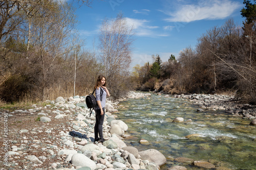 Young beautiful girl on nature with a backpack on the river bank. Good mood on a bright spring day. Against the background of the forest and a beautiful mountain river. Tourism and travel.
