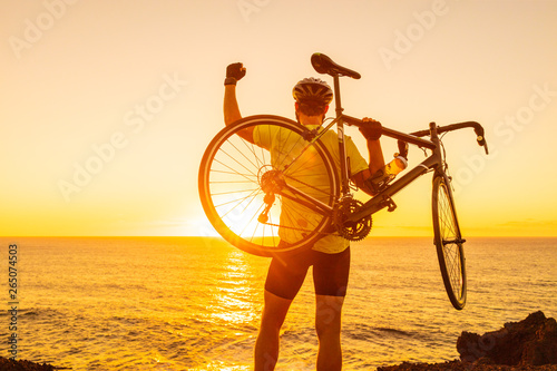 Fototapeta Naklejka Na Ścianę i Meble -  Success, achievement and winning concept with cyclist man road biking. Happy male professional athlete cycling raising arms lifting bike by sea during sunset cheering and celebrating at summit top.