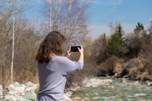 Young beautiful girl on nature takes a selfie on the phone and takes pictures of nature. Against the background of the forest and a beautiful mountain river. Tourism and travel.