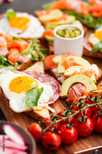 Huge healthy breakfast with sun-dried tomatoes, sandwiches with scrambled eggs, sausage, salmon, arugula, curd cheese, avocado on a wooden board