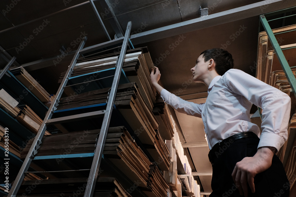 A teenager in a white shirt pulls out a book from the rack in a book depository or archive room. Studying documents. The concept of a historical library, back to school or popularization of reading.