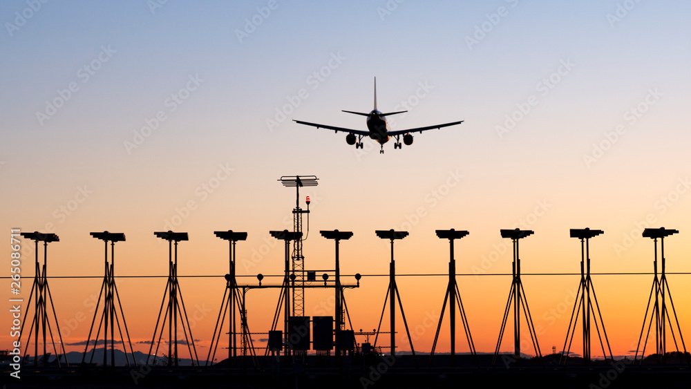 Sunset over an International Airport with the silhouette of a passenger airplane landing on a runway.