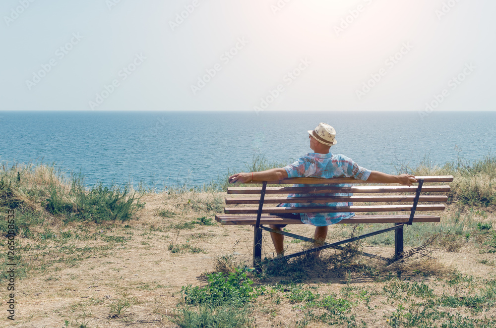 Young man in a hat sits on a bench and enjoys the view of the sea