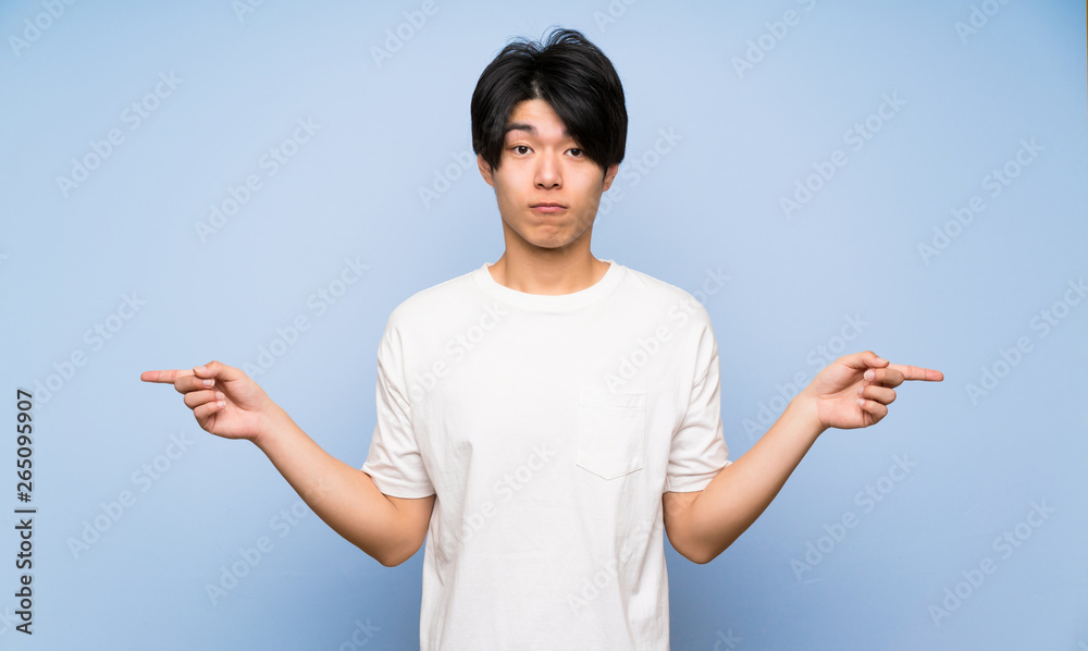 Asian man on isolated blue background pointing to the laterals having doubts Asian man on isolated blue background pointing to the laterals having doubts