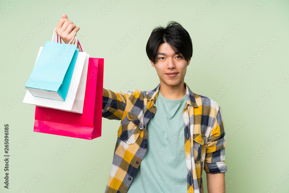 Asian man over isolated green wall holding a lot of shopping bags Asian man over isolated green wall holding a lot of shopping bags