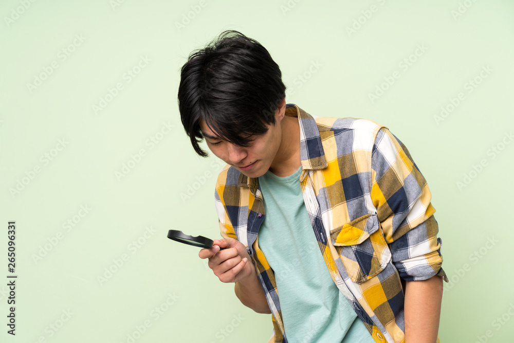 Asian man over isolated green wall holding a magnifying glass