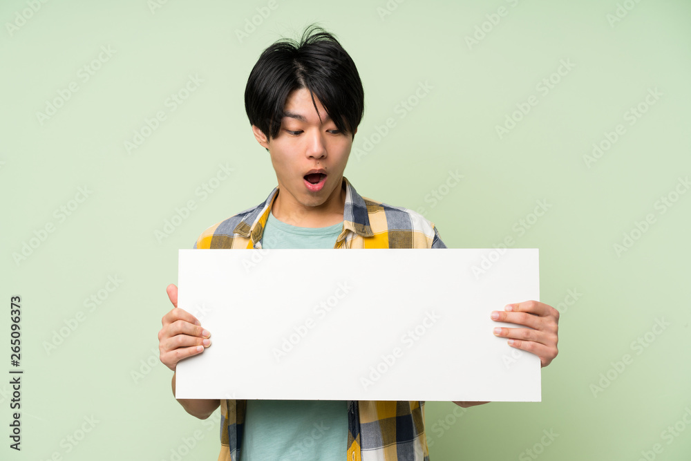 Asian man over isolated green wall holding an empty white placard for insert a concept Asian man over isolated green wall holding an empty white placard for insert a concept