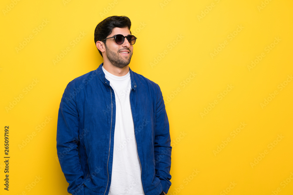 Man with blue jacket over yellow wall with glasses and smiling Man with blue jacket over yellow wall with glasses and smiling