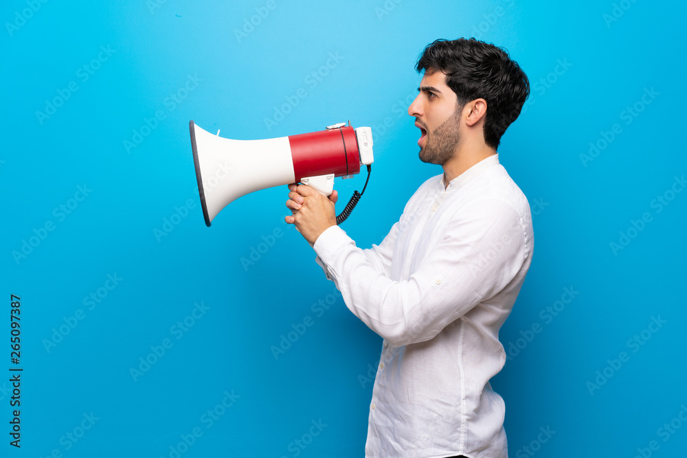 Young man over isolated blue wall shouting through a megaphone Young man over isolated blue wall shouting through a megaphone