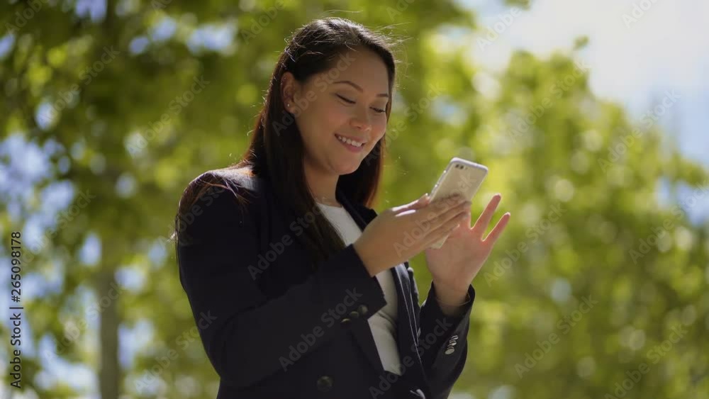 Smiling young woman using smartphone outdoor. Low angle view of beautiful happy girl messaging by cell phone on blurred background. Technology concept