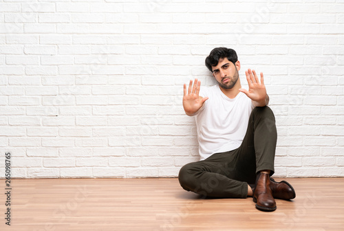 Young man sitting on the floor making stop gesture and disappointed