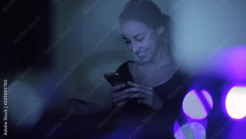 pretty smiling woman sits in the dark room at night, holds a phone in hands, swiping, reading messages. Attractive young Woman Using Mobile Phone at home alone. pretty smiling woman sits in the dark room at night, holds a phone in hands, swiping, reading messages. Attractive young Woman Using Mobile Phone at home alone.