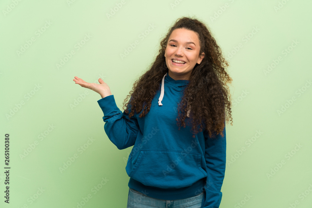 Teenager girl over green wall holding copyspace imaginary on the palm to insert an ad