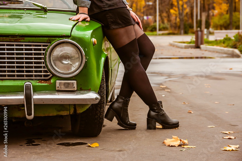 A girl in a denim jacket and black stockings, pantyhose and a black skirt is leaning over the green retro car. Legs closeup