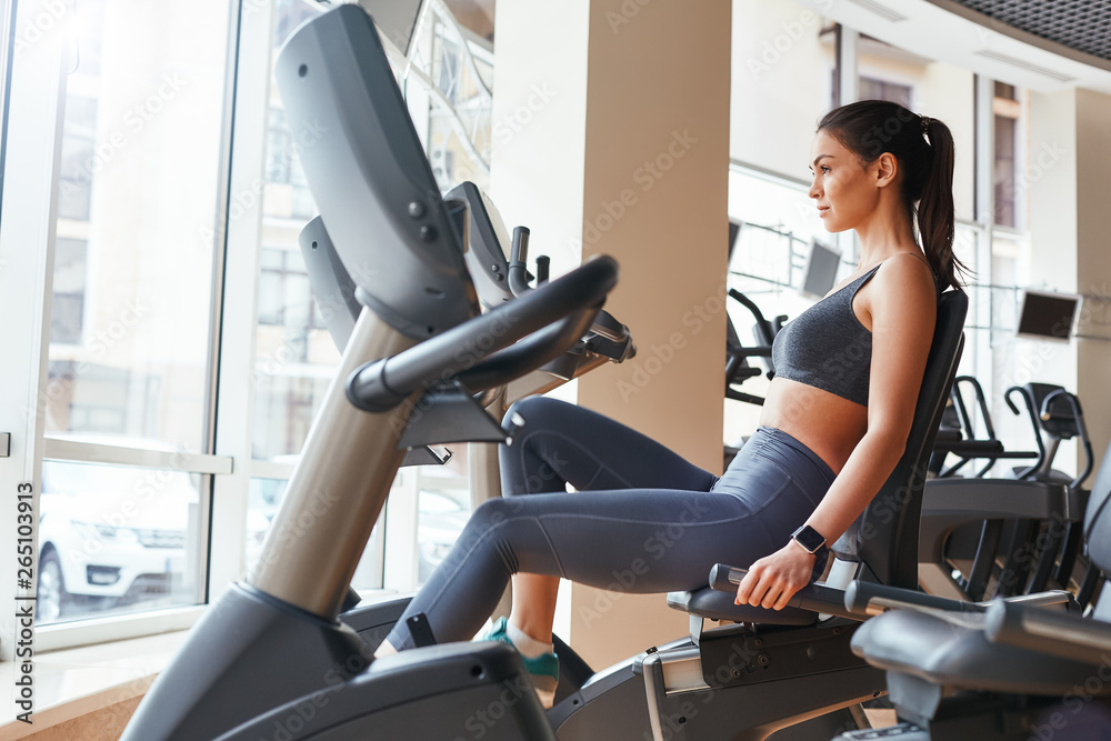 Exercising. Side view of young beautiful woman with perfect body in sportswear and smart watch on her hand is cycling at gym Exercising. Side view of young beautiful woman with perfect body in sportswear and smart watch on her hand is cycling at gym