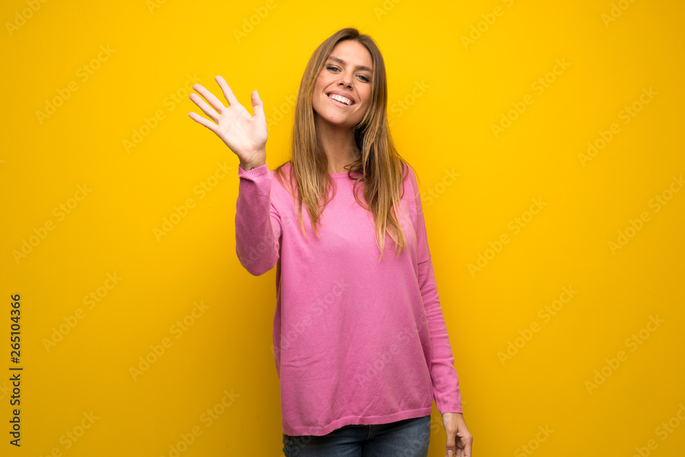 Woman with pink sweater over yellow wall saluting with hand with happy expression Woman with pink sweater over yellow wall saluting with hand with happy expression