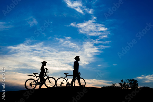 Silhouettes of young couple with bikes at sunset. Girlfriend with boyfriend walking with bicycles on evening sky background. Romantic date on bikes.