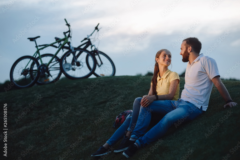Couple of cyclists in love resting on green hill. The pair finished the bike ride on the green meadow. Time spending together is priceless.