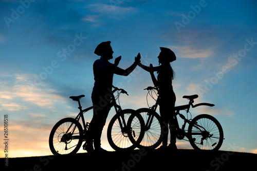 Cheerful couple of bicyclists clapping hands at sunset. Happy young couple having fun riding a bicycle on evening sky background.