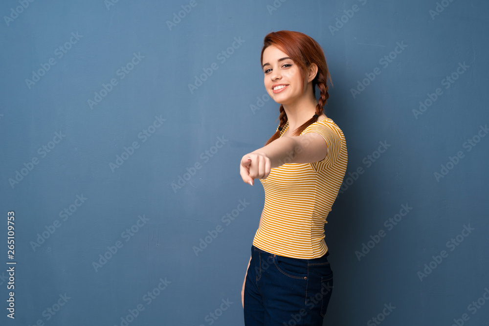 Young redhead woman over blue background points finger at you with a confident expression Young redhead woman over blue background points finger at you with a confident expression