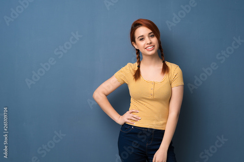 Young redhead woman over blue background posing with arms at hip and smiling © luismolinero