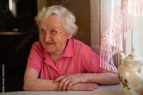 Portrait of an old pensioner woman sitting at a table in the kitchen in her house. Care for lonely elderly.