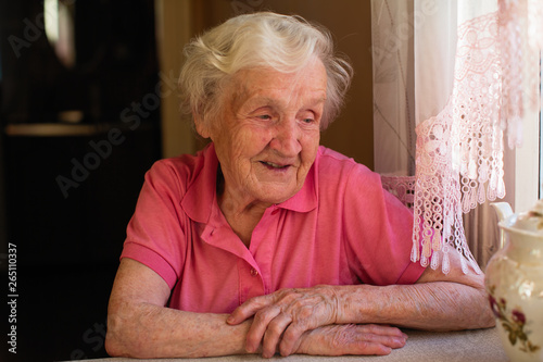 Portrait of an old pensioner woman sitting at a table in the kitchen in her house. Care for lonely elderly.