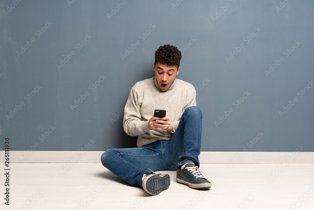 Young man sitting on the floor surprised and sending a message Young man sitting on the floor surprised and sending a message