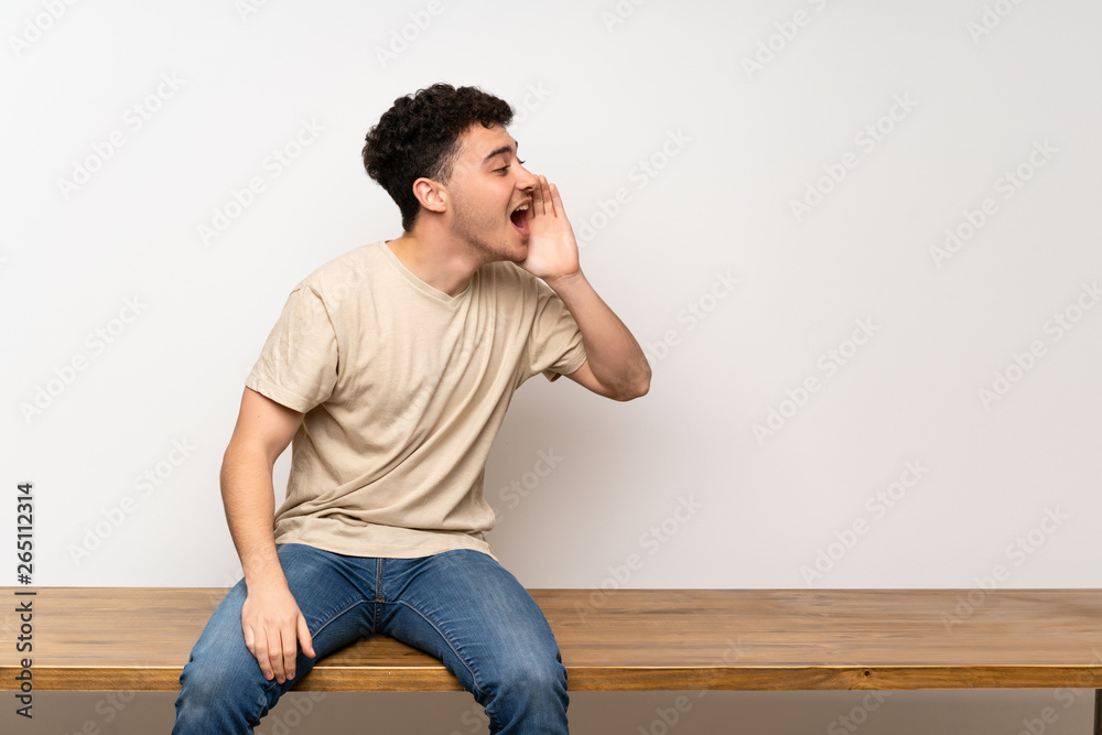 Young man sitting on table shouting with mouth wide open to the lateral Young man sitting on table shouting with mouth wide open to the lateral