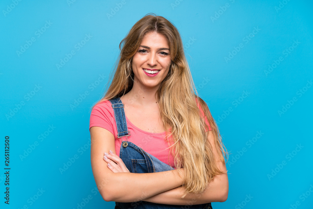Young blonde woman with overalls over isolated blue wall keeping the arms crossed in frontal position Young blonde woman with overalls over isolated blue wall keeping the arms crossed in frontal position