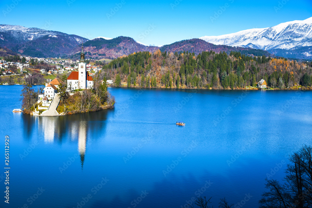 Panoramic view of small natural island in the middle of alpine lake with church dedicated to assumption of Mary and castle with snowy mountain range in the background in winter landscape in Slovenia