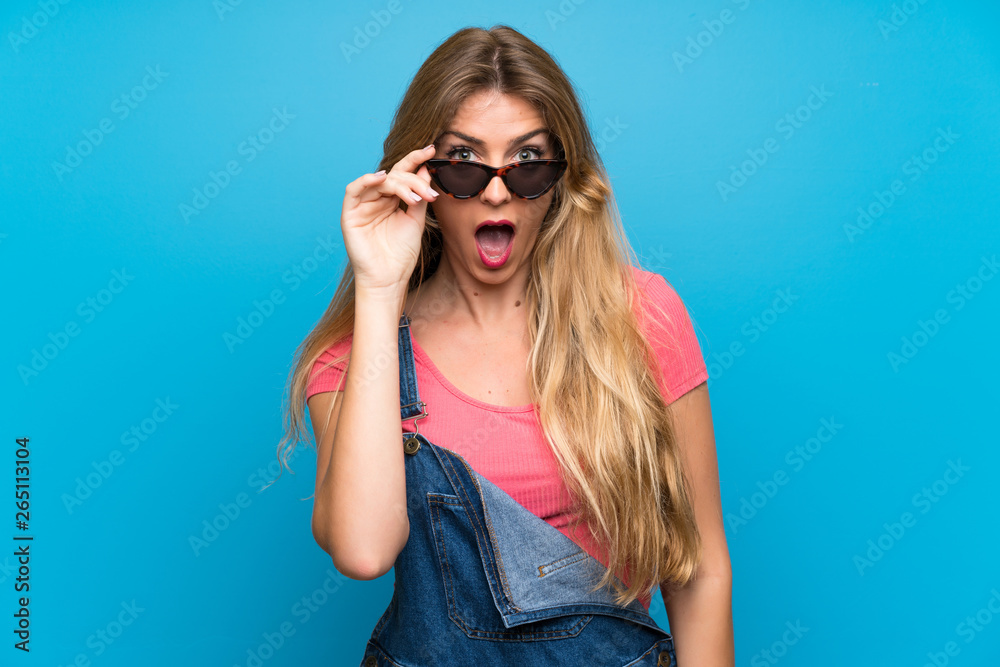 Young blonde woman with overalls over isolated blue wall with glasses and surprised Young blonde woman with overalls over isolated blue wall with glasses and surprised