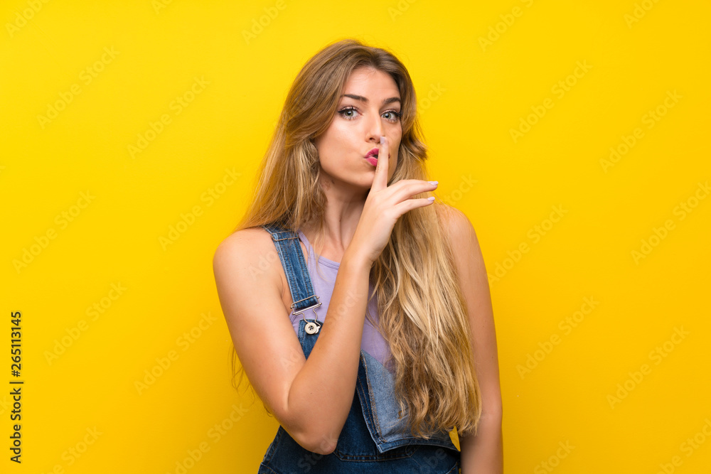 Young blonde woman with overalls over isolated yellow background doing silence gesture Young blonde woman with overalls over isolated yellow background doing silence gesture