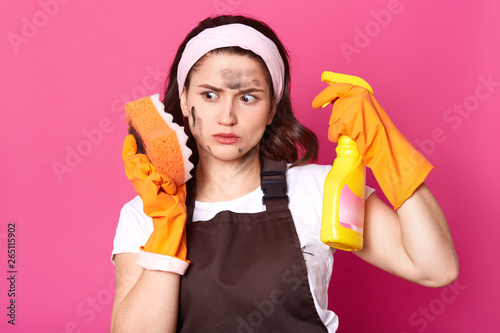 Thoughtful impressed housewife looks at dirty sponge, looks shocked, posing isolated over pink background in studio. Exhausted brunette female wears brown apron, white t shirt and headband, gloves. photo