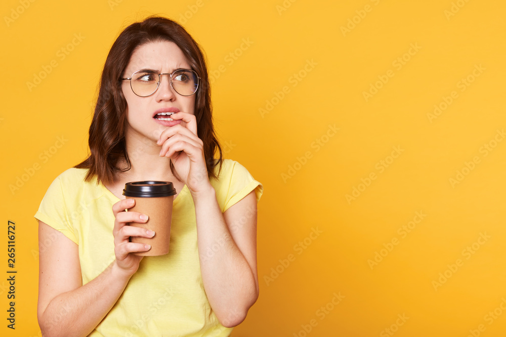 Confused thoughtful young lady looks aside, puts finger in her mouth, holds papercup with coffee in one hand, wearing light yellow casual t shirt and specstacles. Copy space for advertisement. Confused thoughtful young lady looks aside, puts finger in her mouth, holds papercup with coffee in one hand, wearing light yellow casual t shirt and specstacles. Copy space for advertisement.