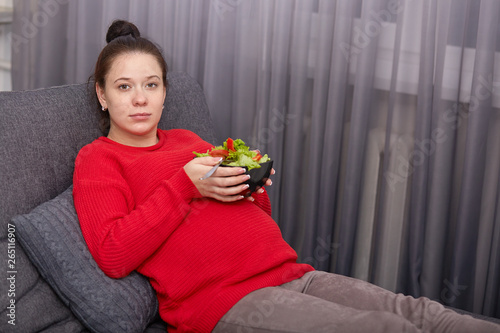Indoor shot of young pregnant woman wears red sweater and maroon leggins  holding bowl of salad in both hands  model looks at camera  sits on gray sofa in living room  having calm expression.