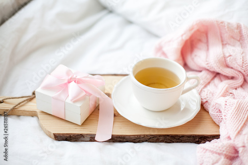 White mug with tea, gift box with ribbon on the bed. Breakfast in bed. Cozy. Pink plaid. Cotton.