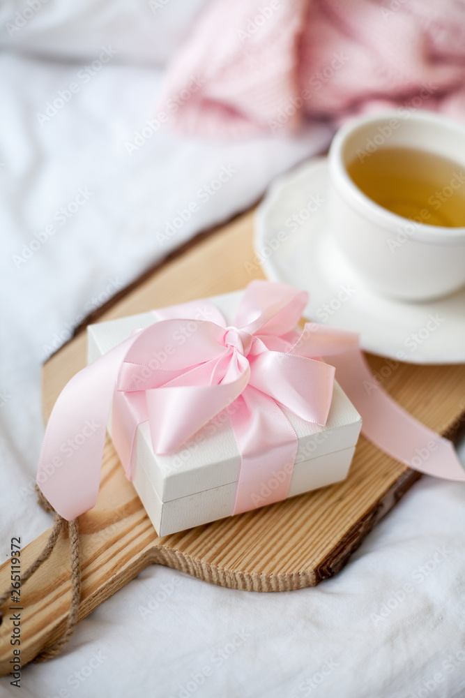 White mug with tea, gift box with ribbon on the bed. Breakfast in bed. Cozy. Pink plaid. Cotton. White mug with tea, gift box with ribbon on the bed. Breakfast in bed. Cozy. Pink plaid. Cotton.