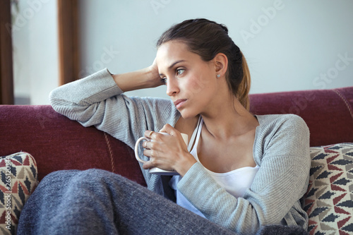 Depressed young woman thinking about her problems while drinking coffee on sofa at home. photo