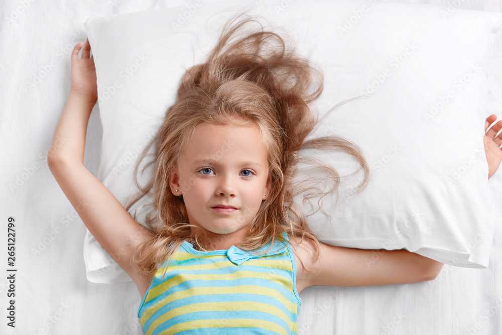 View from above of beautiful small lovely female child with light hair, dressed in striped pyjamass, lies on pillow, has good rest in bed, awakes early in morning. Childhood and leisure concept. View from above of beautiful small lovely female child with light hair, dressed in striped pyjamass, lies on pillow, has good rest in bed, awakes early in morning. Childhood and leisure concept.