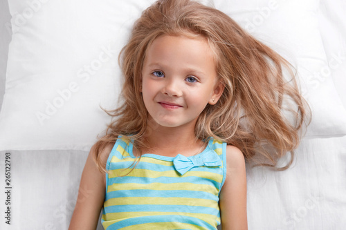 Photo of pleasant looking small female kid with blue eyes, dressed in nightwear, has satisfied expression, lies on white bedclothes, enjoys good rest, rejoices starting new day. Bed time and children