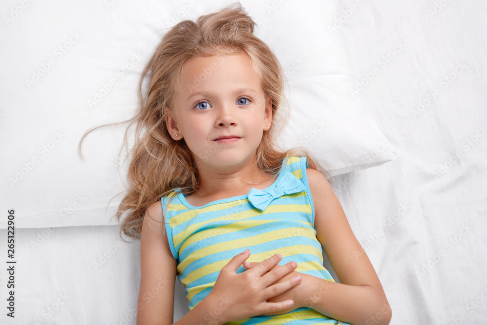 Good looking small kid with pleasant appearance, blue appealing eyes, soft skin, dressed in nightclothes, poses on comfortable bed, listens to mothers story before sleep. Little daughter in bedroom Good looking small kid with pleasant appearance, blue appealing eyes, soft skin, dressed in nightclothes, poses on comfortable bed, listens to mothers story before sleep. Little daughter in bedroom