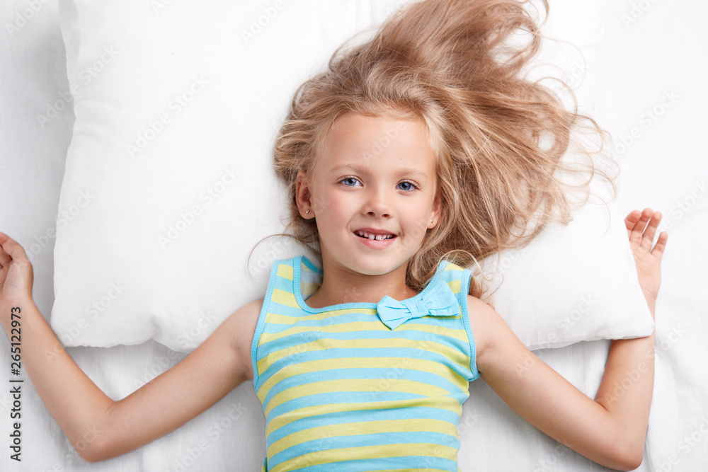 Photo of small cute female child has attractive look, lies in comfortable bed on pillow, dressed in nightwear, enjoys good morning, smiles positively at camera. Children and awakening concept Photo of small cute female child has attractive look, lies in comfortable bed on pillow, dressed in nightwear, enjoys good morning, smiles positively at camera. Children and awakening concept